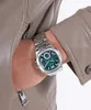 A close-up shot of a man's wrist wearing a silver watch with a green dial, partially tucked into the pocket of a light brown leather jacket. Produktbild-Vorschau