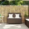 A brown poly rattan garden bench with white cushions is placed on a patio in front of a bamboo fence. Trees and foliage are visible in the background. Produktbild-Vorschau