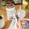 A hand holding art supplies, including pencils and pens, with a plant and art supplies in the background. Produktbild-Vorschau