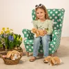 A young girl is sitting in a polka-dotted armchair holding a stuffed bunny. There is a basket of flowers and more stuffed bunnies in front of her. Náhled obrázku produktu