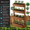 A multi-tiered wooden planter filled with plants, set against a wooden fence and green foliage. A small bird is perched on the fence. Produktbild-Vorschau
