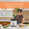 A woman is standing in a kitchen next to a white air fryer, with various prepared meals on the counter. Náhľad obrázku produktu