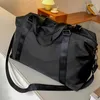 A black duffle bag is placed on a white surface, with a blurred background showing a wooden shelf and a lamp. Voir les images du produit