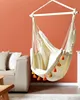 A beige hammock chair with orange tassels hangs in a room with an orange wall and a beige sofa. Produktbild-Vorschau