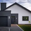 A modern house exterior with a dark gray garage door, entrance door, and chimney, contrasted with a white wall and a dark roof. The driveway is paved with gray bricks. Produktbild-Vorschau