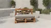 A wooden pallet bench with a gray cushion is placed on a patio next to a small pool and some plants. Produktbild-Vorschau