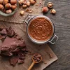 A top-down view of chocolate spread in a jar, surrounded by hazelnuts and chocolate chunks on a wooden table. Produktbild-Vorschau