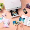 A Canon PIXMA printer is printing photos, with printed photos, a smartphone, and a notebook on a wooden desk. Podgląd zdjęcia produktu