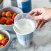 A hand is shown lifting a fresh lid from a container of yogurt, with bowls of fruit in the background. Anteprima dell’immagine prodotto