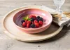 A pink bowl filled with raspberries and blackberries sits on a wooden tray, with a napkin and glass in the background. Náhled obrázku produktu