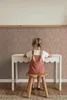 A young girl sits at a small white table, facing away from the camera, with her back to the viewer. She is wearing a terracotta-colored overall dress and a white t-shirt. The wall behind her has a speckled pink and brown wallpaper. The floor is wooden with Podgląd zdjęcia produktu