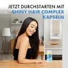 A woman with long, dark hair is smiling and touching her hair, with a bottle of Shiny Hair Complex Kapseln on a wooden table in the foreground. Produktbild-Vorschau