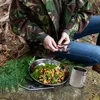 A person is sitting outdoors in a forest setting, preparing to eat a meal from a camping dish. There is a metal mug and cutlery on a log next to the dish. Produktbild-Vorschau