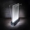 A product shot of a clear acrylic LED table stand with a black base, set against a dark, textured background. Náhľad obrázku produktu