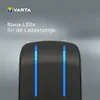 A close-up of a black charging device with two bright blue LED lights indicating charging status, set against a gradient gray background. The VARTA logo and German text are visible in the upper left corner. Voir les images du produit