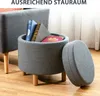 A round storage ottoman with its lid open, revealing books inside, placed on a wooden floor next to another piece of furniture. Produktbild-Vorschau