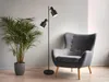A black floor lamp with two adjustable spotlights stands next to a gray armchair and a large potted plant in a modern living room. Produktbild-Vorschau