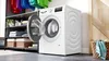 A white Bosch Serie 4 washing machine with its door open, placed in a laundry room next to shelves with folded laundry and baskets, and a door in the background. Produktbild-Vorschau