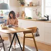 A woman is sitting at a kitchen table reading a book and holding a mug, with a kitchen counter and sink in the background. Produktbild-Vorschau