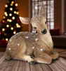 A decorative deer figurine covered in artificial snow, placed on a wooden surface in front of a blurred Christmas tree and window. Produktbild-Vorschau