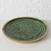 A green ceramic plate with a carved pattern on a wooden base, placed on a white surface in front of a white brick wall. Produktbild-Vorschau