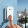 A finger presses a wireless doorbell mounted on a blue wall, with a blurred cityscape in the background. Náhľad obrázku produktu