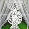 A white sheer curtain with decorative cutouts hangs in front of a window with a green outdoor view. Podgląd zdjęcia produktu