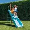 A young girl slides down a blue slide in a backyard, with a woman standing behind her. Náhled obrázku produktu