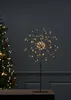 A starburst-shaped LED light decoration stands on a dark surface, with a blurred Christmas tree in the background. Produktbild-Vorschau
