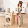 A young girl is playing with a wooden toy washing machine, laundry basket, and ironing board in a bright, domestic setting. Produktbild-Vorschau