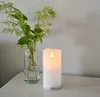 A white LED candle sits on a white surface next to a glass vase with green foliage. Produktbild-Vorschau