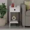 A modern white nightstand with a drawer and shelf, placed next to a sofa. Decorated with vases and books, and a potted plant nearby. Podgląd zdjęcia produktu