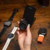 A hand holding a black USB-C charging adapter for a smartwatch, with other smartwatches and a charging stand visible on a wooden surface. Produktbild-Vorschau