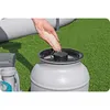 A hand is pouring sand into a sand filter for a swimming pool, which is set up on artificial grass. Produktbild-Vorschau