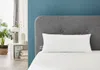A white pillow is placed on a bed with a grey headboard against a blue wall. A plant and a side table are visible on the left. Produktbild-Vorschau