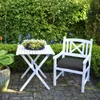 A white garden table and chair set with a plant on the table, placed on a paved patio surrounded by lush green bushes and trees. Náhľad obrázku produktu