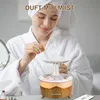 A woman in a bathrobe and towel is adding essential oil to a mushroom-shaped humidifier that is emitting mist and has a warm light. Produktbild-Vorschau