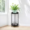 A black metal corner plant stand with two shelves, holding a green potted plant on the top shelf and two books on the bottom shelf, placed in a corner near a window with sunlight casting shadows on the wall. Podgląd zdjęcia produktu
