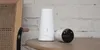 A white tower-like device and a white cylindrical camera are placed on a wooden table next to a wooden chair. Produktbild-Vorschau
