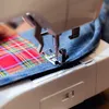 A close-up shot of a sewing machine needle and presser foot stitching together two pieces of fabric: one denim and one plaid. Produktbild-Vorschau