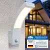 A white LED outdoor light with a motion sensor is mounted on a textured wall, with a blurred house in the background. There are also several certification badges and text overlays. Produktbild-Vorschau