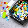 A collection of colorful building blocks scattered on a gray, fuzzy surface, with a black storage bin filled with more blocks. Anteprima dell’immagine prodotto