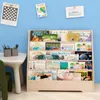 A wooden bookshelf filled with children's books is placed against a light blue wall. There are also some framed pictures and a small table with art supplies on the left side of the image. Produktbild-Vorschau