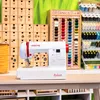 A white sewing machine with red accents is displayed on a wooden table, surrounded by various sewing supplies like spools of thread and buttons. Produktbild-Vorschau