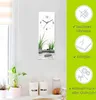 A vertical wall clock with a zen-like image of stones and grass in water, displayed on a white wall next to kitchen canisters. Produktbild-Vorschau