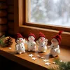 Four decorative snowmen with red hats are arranged on a wooden windowsill, with a snowy window in the background and pine sprigs and stars scattered around them. Produktbild-Vorschau