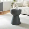 A dark gray concrete side table with a textured finish is placed on a white rug next to a sofa. A white mug and a magazine are on top of the table. Anteprima dell’immagine prodotto