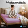 A young boy sitting cross-legged on a pink fitted sheet on a bed, with pillows and a bedside table with a lamp and mug. Produktbild-Vorschau
