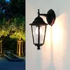 An outdoor wall lamp is mounted on a white wall, with a garden scene visible on the left. Podgląd zdjęcia produktu