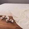 A cream-colored fitted sheet is draped over a mattress, with cotton branches on a wooden floor in the foreground. Náhled obrázku produktu